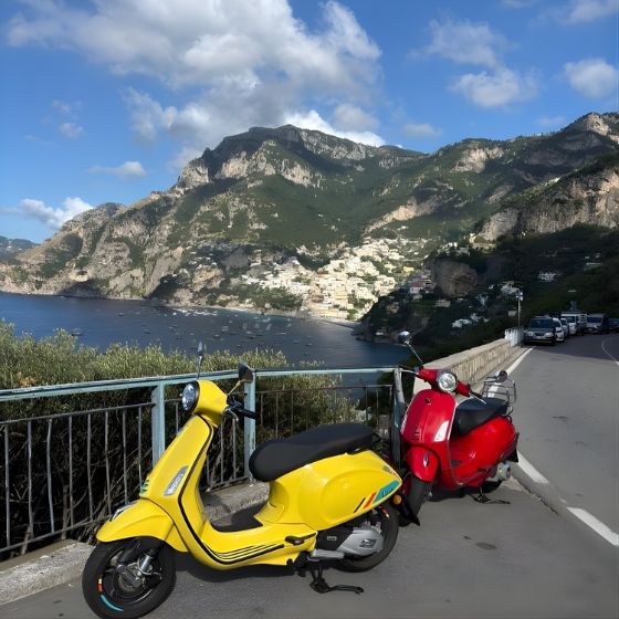 A yellow and a red Vespa parked on a scenic coastal road with a breathtaking panoramic view of Positano and the sea.